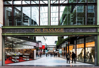 Entrance to De Passage shopping arcade with glass roof, stores on both sides and people walking inside.