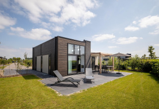 Lodge moderna a Holiday Park «Zeedijk 2» nei Paesi Bassi con patio, lettini prendisole e giardino erboso.