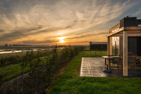 Solnedgang ved en moderne lodge med terrasse, udsigt over marker, flod og græssende køer i det fjerne.