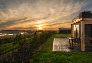 Sonnenuntergang bei einer modernen Lodge mit Terrasse, Blick auf Felder, Fluss und grasende Kühe.