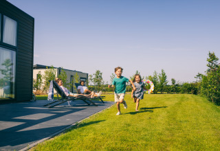 Two children run on the grass at a lodge while two adults relax on lounge chairs on the patio.