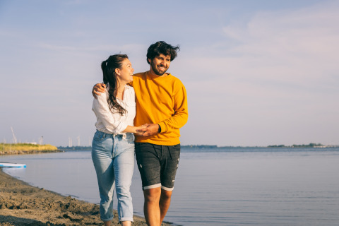 Et ungt par går smilende langs stranden ved en lodge med armene omkring hinanden på en solrig dag.