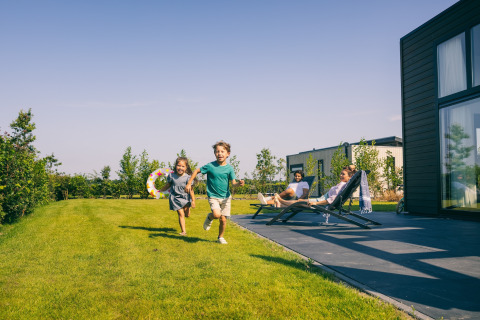 Children run across a grassy lawn at Holiday Park Zeedijk 2, Netherlands, while adults relax by the lodge.