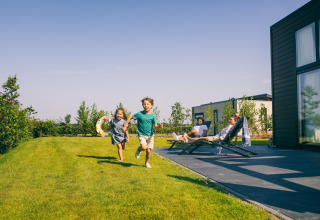 Kinder rennen über die grüne Wiese vor einer modernen Lodge, Erwachsene entspannen sich auf Liegestühlen.