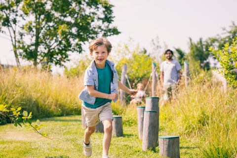 Un bambino felice corre all'aperto su un parco giochi naturale in un lodge, con la famiglia sullo sfondo.