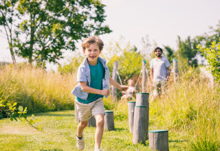 En glad dreng løber udendørs på en naturlegeplads ved en lodge, med familie i baggrunden.