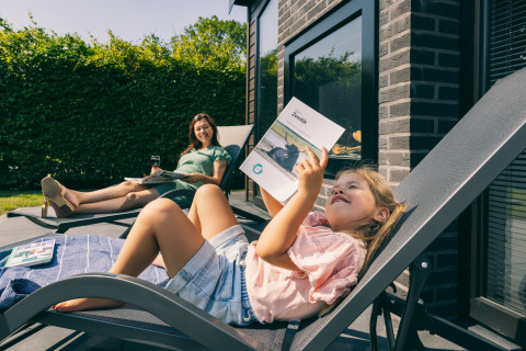 Pige og kvinde slapper af i solen på liggestole ved en lodge, mens de læser og nyder deres tid sammen.