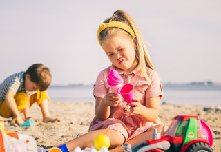 Zwei Kinder spielen am Sandstrand mit Spielzeug, ein Mädchen verzieht das Gesicht, während ein Junge schaufelt.