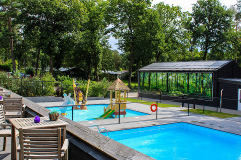 Área de glamping al aire libre con piscinas, zona de juegos infantiles y árboles verdes en un día soleado.