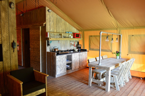 Interior of a safari tent featuring a small kitchen, wooden table with chairs, and cozy lighting.