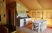 Interno di una tenda safari con cucina attrezzata, tavolo in legno, sedie e illuminazione calda.