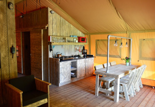 Interior of a safari tent featuring a small kitchen, wooden table with chairs, and cozy lighting.