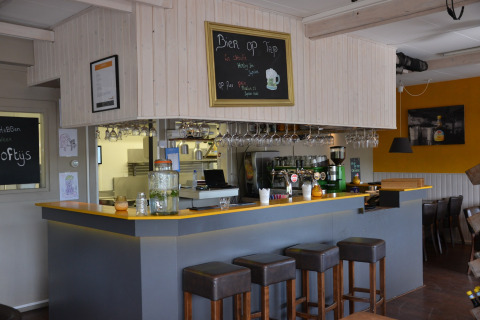 Photo of a cozy bar setup inside a safari tent, featuring stools, a counter, hanging glasses, and a menu board.