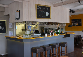 Photo of a cozy bar setup inside a safari tent, featuring stools, a counter, hanging glasses, and a menu board.