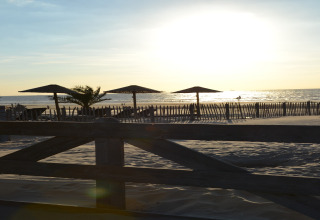 Sunset view from a safari tent at the beach, showing parasols, sandy dunes, and a wooden fence in front.