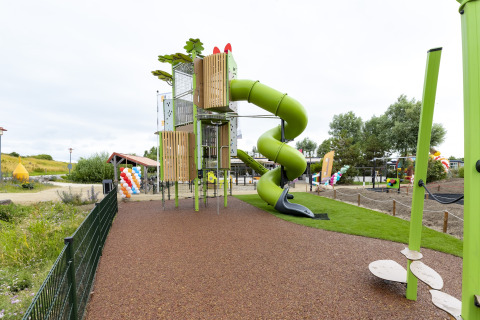 Playground with a green spiral slide at a safari tent, fenced and with colorful balloons in the background.