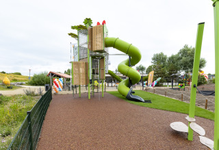Parque infantil con tobogán verde en espiral junto a una tienda safari, cercado y globos de colores al fondo.