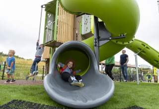 Children playing on a playground with a slide and climbing equipment while adults watch nearby.