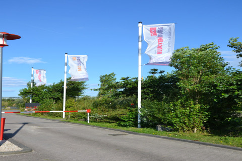 Entrance to safari tent area with fluttering flags, lush greenery, and a barrier on a sunny day.