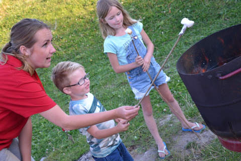 A woman and two children toast marshmallows over a fire barrel near a safari tent on green grass.