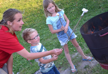 Una donna e due bambini tostano marshmallow su un fuoco vicino a una tenda safari su erba verde.