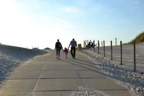 A family walks hand in hand along a paved path by the sandy beach during a bright, sunny day.