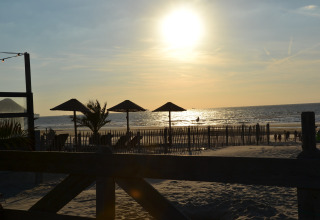 Sunset view from a safari tent terrace facing the beach, with straw umbrellas and lounge chairs in sand.