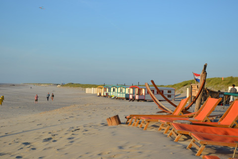 Beach scene with empty deck chairs, colorful beach huts, and people walking along the sandy shore at sunset.