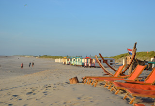 Beach scene with empty deck chairs, colorful beach huts, and people walking along the sandy shore at sunset.