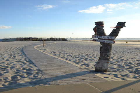 Sandy beach with a wooden path, a V-shaped post with signs, and sunny weather with huts in background.