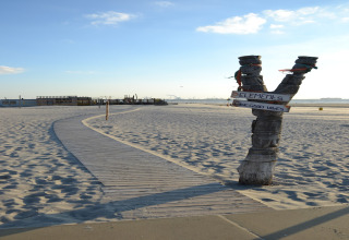 Sandy beach with a wooden path, a V-shaped post with signs, and sunny weather with huts in background.