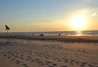 Sunset over a serene sandy beach with footprints, people by the water, and a safari tent marker left.