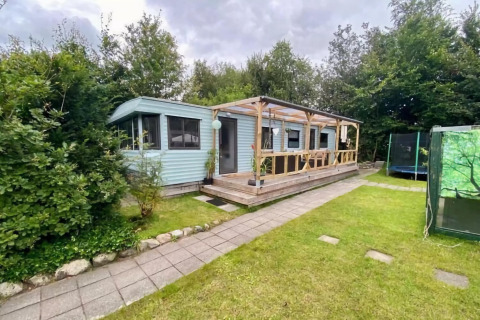 Mobilhjemmet Sunhouse Bodhi med overdækket terrasse, grøn have og omgivet af træer under overskyet himmel.