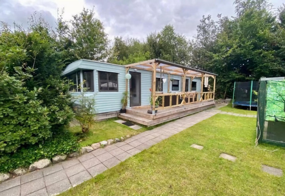 Mobilhjemmet Sunhouse Bodhi med overdækket terrasse, grøn have og omgivet af træer under overskyet himmel.
