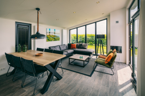 Moderne opholdsstue på Holiday Park «Zeedijk 3» i Holland med store vinduer, spisebord og sofa.