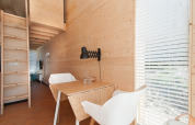 Interiør af et moderne tiny house med trævægge, spisebord, hvide stole og store vinduer med persienner.