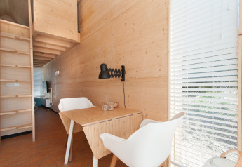 Interiør af et moderne tiny house med trævægge, spisebord, hvide stole og store vinduer med persienner.