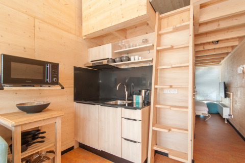 Cuisine moderne de tiny house avec murs en bois, échelle vers mezzanine et chambre visible derrière.