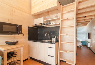 Cuisine moderne de tiny house avec murs en bois, échelle vers mezzanine et chambre visible derrière.