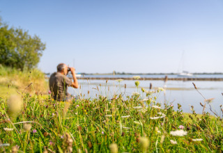 Homme aux jumelles près du lac à Eco Resort Grevelingenstrand, entouré de fleurs sauvages et herbes.