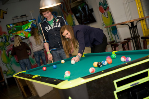 Twee jongeren spelen poolbiljart in een kleurrijke lodge met graffiti op de muren en gasten op de achtergrond.