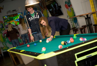 Twee jongeren spelen poolbiljart in een kleurrijke lodge met graffiti op de muren en gasten op de achtergrond.