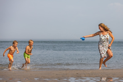 A woman in a floral dress playfully squirts water at two boys running on a Dutch beach by the sea.
