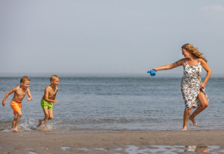 A woman in a floral dress playfully squirts water at two boys running on a Dutch beach by the sea.