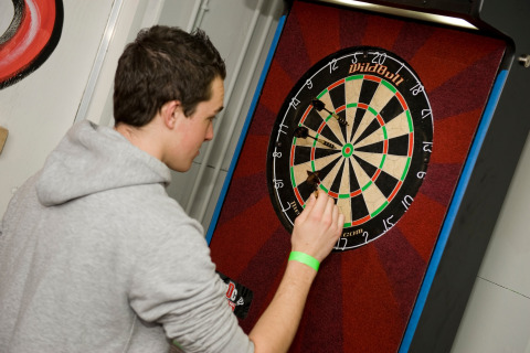 Een man in een grijze hoodie haalt darts uit een dartbord in een lodge, gefocust op het dartspel.