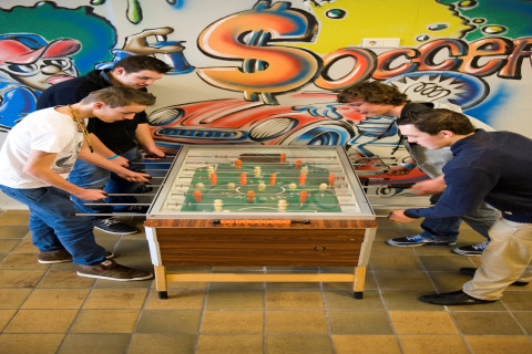 Vier jongeren spelen tafelvoetbal in een lodge, kleurrijke graffitiwand met het woord 'Soccer'.