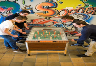 Vier jongeren spelen tafelvoetbal in een lodge, kleurrijke graffitiwand met het woord 'Soccer'.