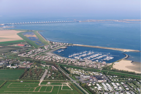 Luchtfoto van een jachthaven met boten, lodges, landbouwgrond en een kustdam op de achtergrond.