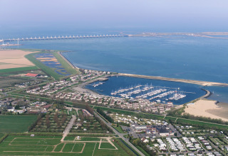 Luchtfoto van een jachthaven met boten, lodges, landbouwgrond en een kustdam op de achtergrond.