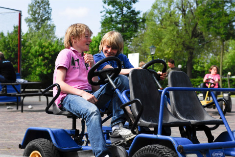 Twee jongens maken plezier op een blauwe skelter bij een lodge, met bomen en mensen op de achtergrond.